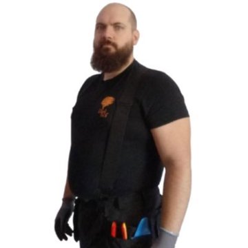 Photo of a bearded, muscular man wearing a dark grey t-shirt and work overalls with tools in the pockets, standing against a white background.