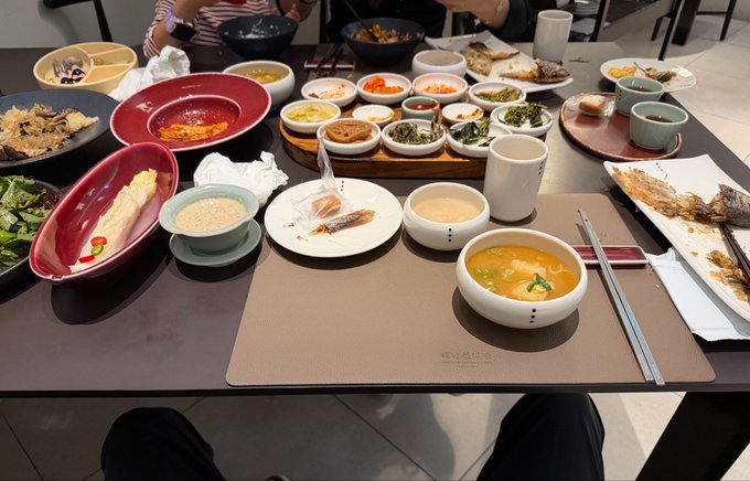 A first-person view of a Korean dining table filled with numerous small banchan dishes, soups, grilled fish, and bowls set on a placemat, showing the abundant spread implied by the caption about ordering dozens of dishes at a Seoul restaurant for 30,000 won.
