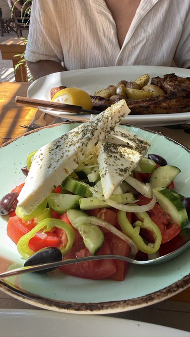 A Greek salad with feta cheese, tomatoes, cucumbers, peppers, and olives sits in the foreground, with grilled fish and potatoes visible on another plate behind it.