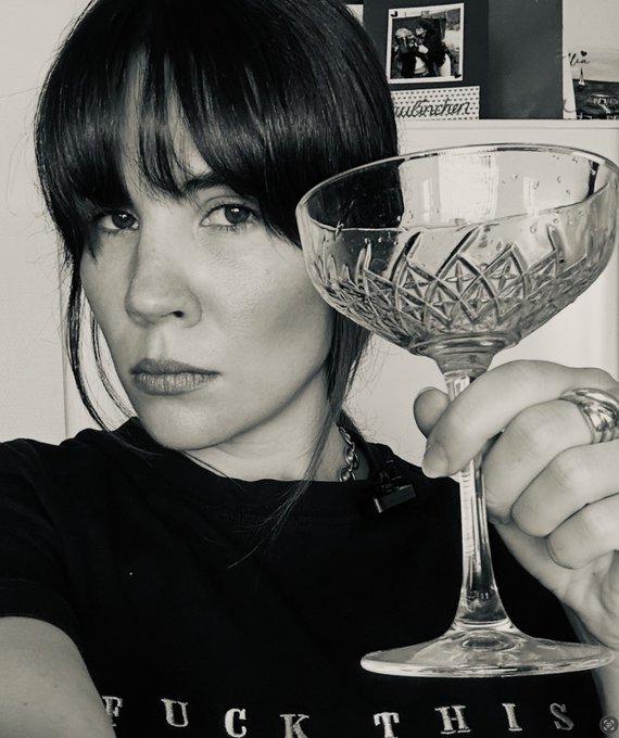 A black-and-white selfie of a woman with dark bangs holding up a crystal coupe glass, wearing a shirt that reads 'FUCK THIS'.