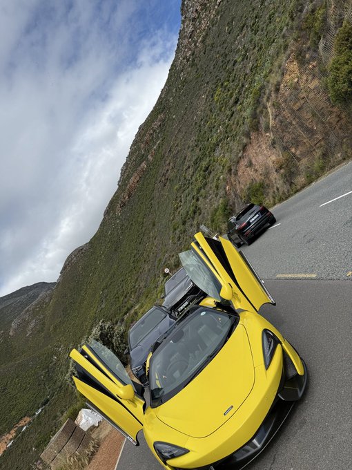 A yellow McLaren sports car with its doors open sits on a mountain road next to a dark SUV, with steep hillsides in the background.