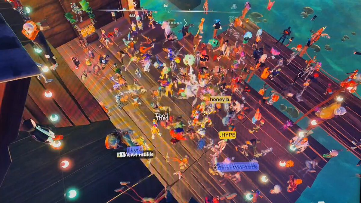 The image shows a large group of people gathered together, possibly at an event or party. The crowd consists of various individuals standing around each other, creating a lively atmosphere. There are also signs posted throughout the scene, which may provide information about the event or location. The overall setting appears to be indoors with a wooden floor and a blue wall in the background.