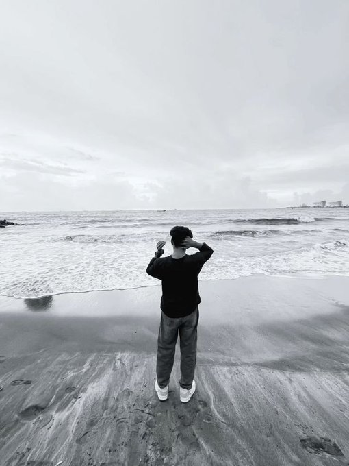 A black-and-white photo of a person standing on a sandy beach, facing the ocean with their hands raised near their face.