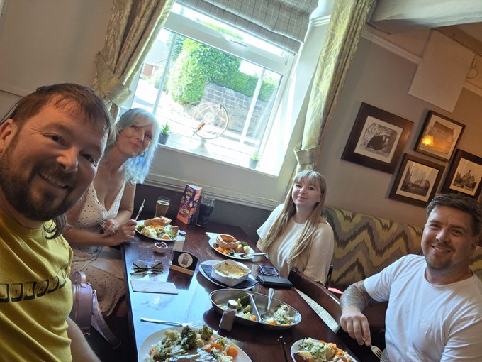 Four people are gathered around a table enjoying a hearty roast beef dinner at what appears to be a cozy pub or restaurant, with framed pictures on the walls and a window showing greenery outside. The image matches the post by showing a satisfying meal, complementing the "smashed a roast beef dinner" caption alongside the playful "Thanks XRP" sign-off.