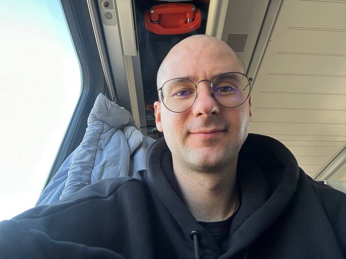 A bald man with round glasses wearing a black hoodie taking a selfie on a train, with a window, jacket, and luggage visible behind him.