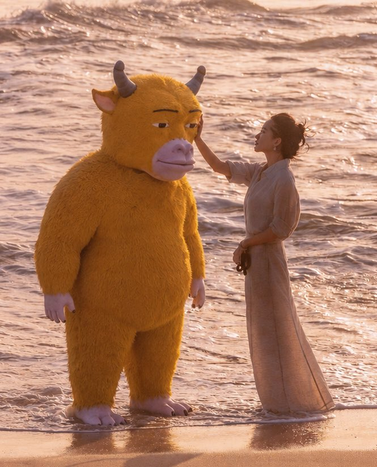 A furry yellow bull creature with horns stands in the ocean surf next to a woman in a flowing beige dress who gently touches its face, evoking a surreal, fairytale-like bond. The image visually captures the "bull is coming" sentiment in the caption, with the literal horned bull symbolizing an approaching bullish market surge.