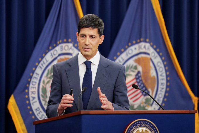 A Federal Reserve official stands at a podium bearing the seal of the Board of Governors, with two Fed flags behind him, as he addresses the recent FOMC decision to hike rates by 25 basis points—the first increase since July 2023—approved unanimously by all 12 members.