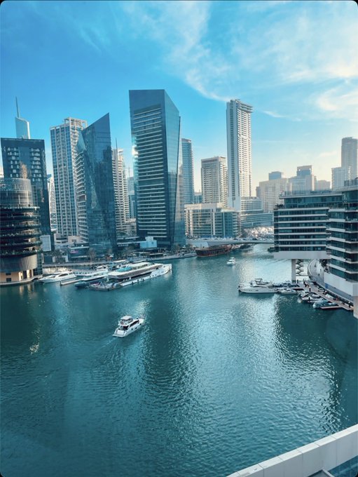 An aerial view of a modern marina cityscape, likely Dubai Marina, with tall skyscrapers, luxury yachts, and boats docked along turquoise waterways under a blue sky.