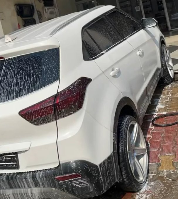 A white SUV is being washed in a car wash bay, with soap suds dripping down its rear and side panels; the vehicle's license plate is blurred for privacy.