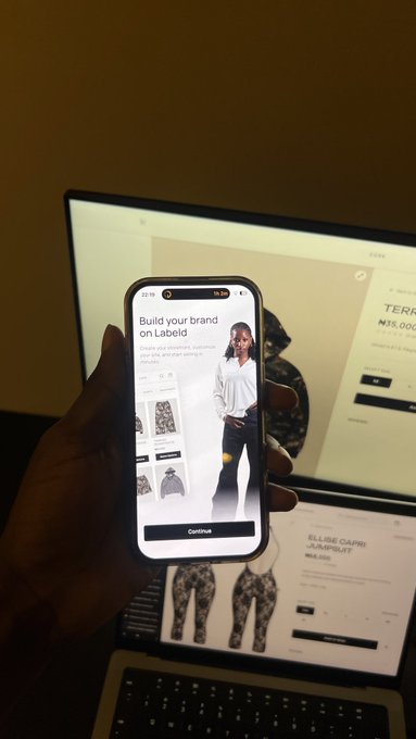 A hand holds a smartphone displaying the 'Build your brand on Labell' app onboarding screen featuring a model, with a laptop showing a Nigerian e-commerce store in the background.