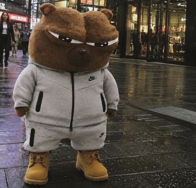 A grumpy-looking brown bear mascot wearing a gray Nike tracksuit and tan Timberland boots, standing on a wet city street.