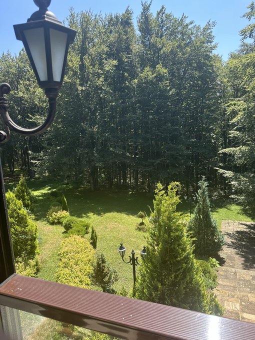 A view from a balcony looking out over a lush green garden with trees and shrubs, featuring an ornate black lamp in the foreground.