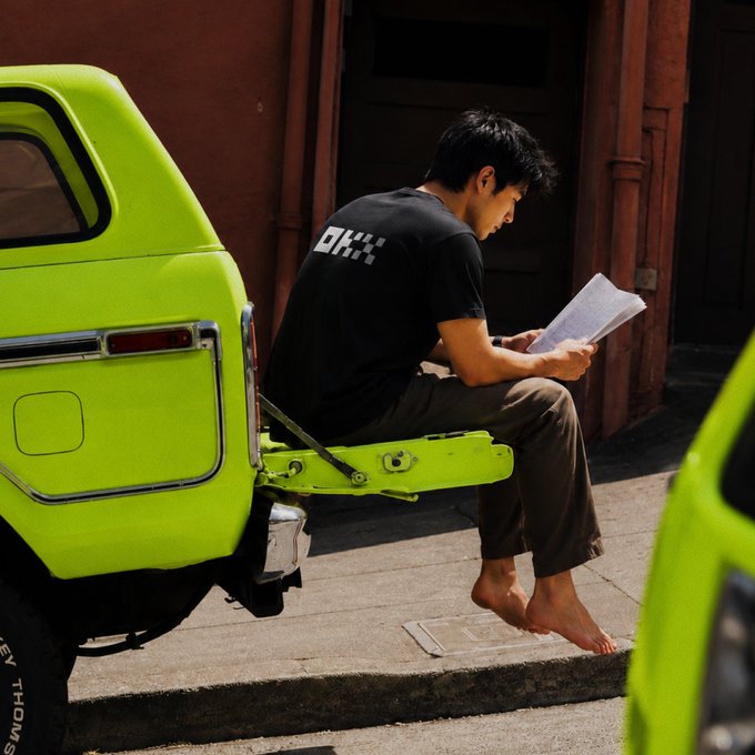 A barefoot man in a black t-shirt and green pants sitting on the open tailgate of a bright green truck, reading a document on a sunny street.