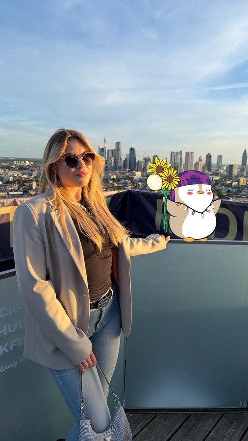 A blonde woman in a beige blazer and sunglasses poses on a rooftop with a city skyline behind her, holding out her hand toward a cute cartoon penguin sticker wearing a purple beanie and holding sunflowers. Partially visible text reads "CK O," "HUD," and "KFU," likely referencing the web3 community mentioned in the post, where she appears to be celebrating her onboarding with her new "Lil" companion.