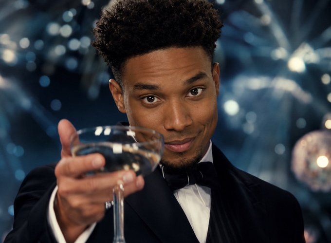 A man in a tuxedo with a bow tie holds up a glass of clear liquid toward the camera, set against a bokeh blue background with sparkling lights.