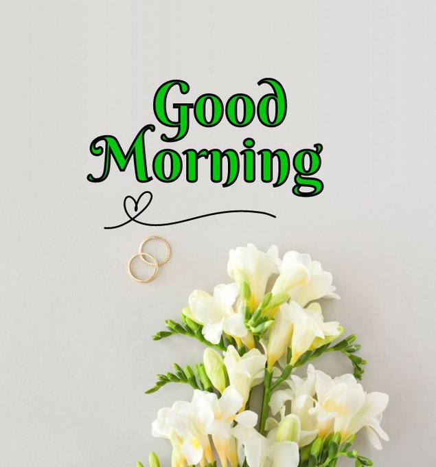 The image features a bouquet of white flowers, possibly roses or lilies, placed on a table against a gray background. The text "Good morning" is written above the flowers, indicating that it's a good morning greeting. There are also two gold rings visible in the scene, which could be part of an engagement or wedding theme for the post.