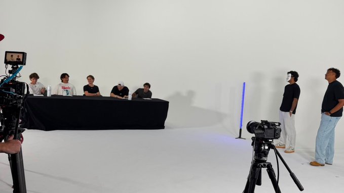 The image shows a film/photo shoot in a white studio, with five people seated at a table on the left facing two standing individuals on the right, all being recorded by cameras. The setup appears to be preparing content for a crypto-focused media production, aligning with the post's message about making crypto content less niche and boring by creating more engaging, visual material.