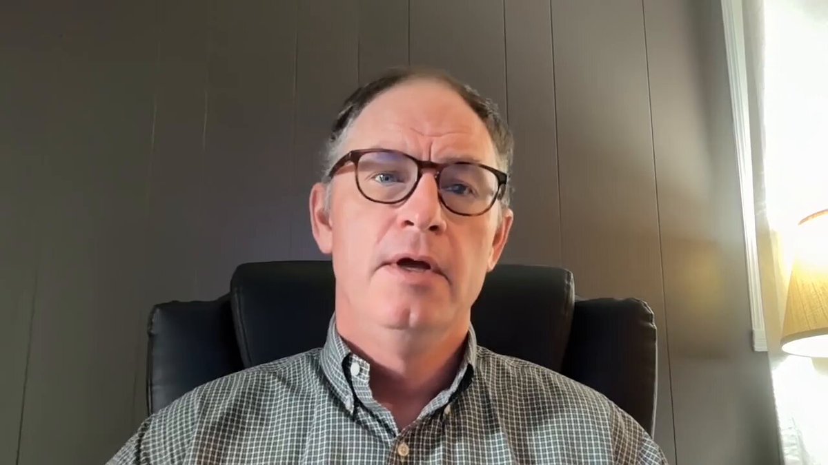 The image shows a man wearing glasses sitting in an office chair, with his face turned towards the camera. He appears to be talking or giving a presentation, as indicated by him looking at the viewer directly. The background of the image features a gray wall, which contrasts with the white lamp that is visible behind the man. There are no texts present in the image, and the focus remains on the main subject - the man sitting in an office chair.