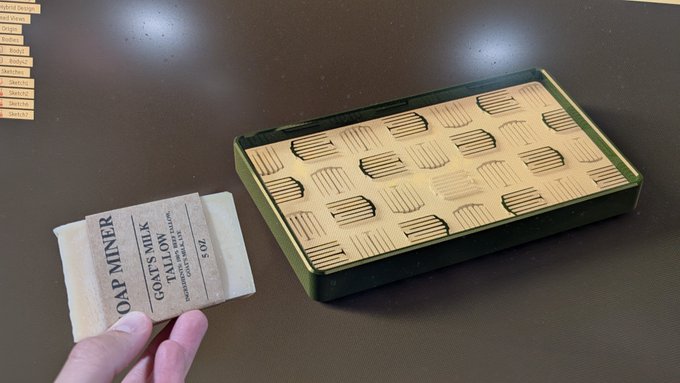 A hand holding a bar of 'Oat Miner Goat's Milk Lavender' soap next to an open green box lined with ridged cardboard on a brown surface.