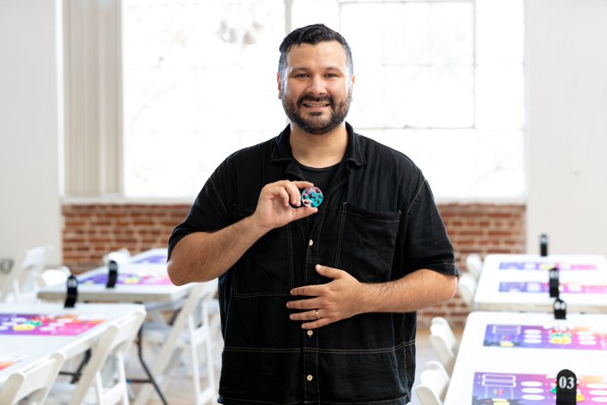 A bearded man in a black shirt smiles while holding a small blue object in a room with tables and large windows.