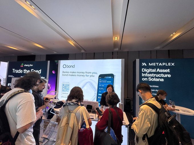 An indoor trade show or conference scene with booths for crypto/Web3 companies, including 'cherryfun' with 'Trade the Food', 'bond' displaying a phone interface and QR code with text 'Banks make money from you, bond makes money for you', and 'METAPLEX Digital Asset Infrastructure on Solana'.