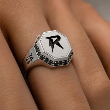 A close-up of a silver ring with a hexagonal face bearing a black 'R' logo, worn on a person's finger.