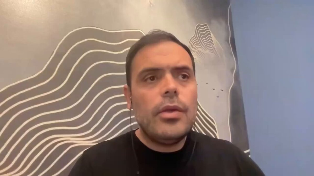 The image shows a man wearing a black shirt, sitting down with his head tilted back as if he's listening intently or looking off into space. The background features a blue wall with white lines that create an abstract pattern. There are also two birds in the image, one on top of the other, which adds to the overall composition and atmosphere of the scene.