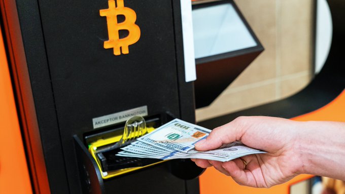 A hand inserts U.S. currency into an orange Bitcoin ATM marked with the iconic "₿" logo and an "ACCEPTOR BANKNOTE" slot, illustrating the unregulated crypto exchange infrastructure at the center of the post's critique comparing Arizona's consumer-friendly refund policy to India's high-tax, low-protection crypto environment.