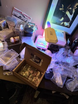 A cluttered desk surface holding a wooden box filled with small items, plastic storage containers, various supplies, a cardboard takeout box, and a monitor displaying an animated character.