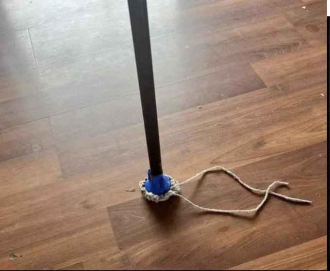 A black pole stands vertically on a dark wood floor, with a blue circular base wrapped in a white rope or crocheted material, and additional loose rope lying on the floor beside it.
