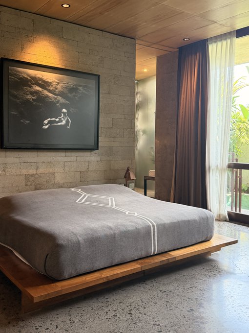 A modern, minimalist bedroom interior features a low wooden platform bed with a gray bedspread featuring white geometric line patterns. A framed black-and-white photograph of a figure in a landscape hangs on a textured stone wall, with floor-to-ceiling curtains and a balcony view visible to the right.