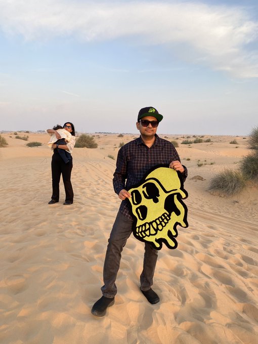 The image features a man holding up a skull-shaped piece of paper art, possibly a painting or a drawing, while standing in a desert setting with sand dunes around him. The scene also includes another person who appears to be taking a picture of the artwork. The caption "Sometimes you just know" suggests that there might be an underlying message or meaning behind the artwork and its display in this particular location.