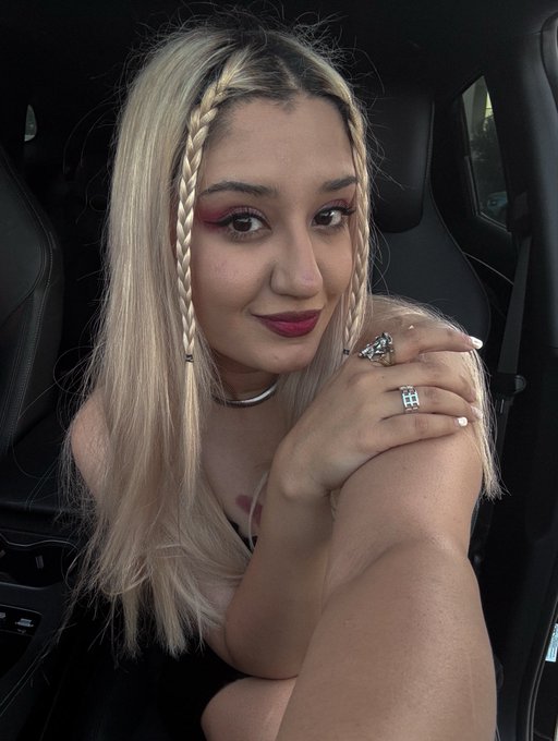 Portrait of a young woman with long blonde hair styled with face-framing braids, wearing pink eyeshadow, sitting in a car.