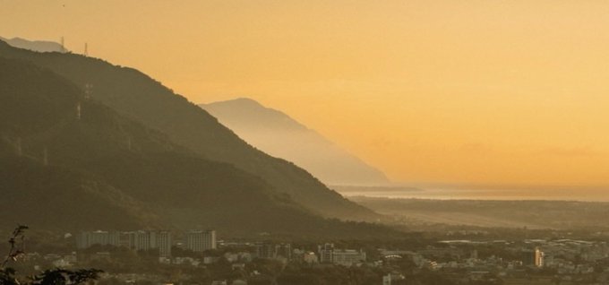 A hazy landscape photograph showing mountains silhouetted against a golden-orange sunset sky, with a cityscape visible in the valley below.