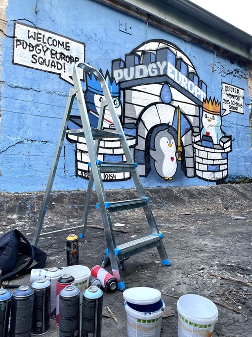A street art mural on a blue wall features penguin characters under a banner reading 'PUDGY EUROPP' with signs stating 'WELCOME PUDGY EUROPP SQUAD!' and 'OCTOBER INVASION 2025 COMING SOON!', with a stepladder and spray paint cans arranged in the foreground.