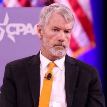 A gray-haired man in a black suit and orange tie speaks at a CPAC event, pictured in the context of breaking news about Michael Saylor's Strategy ($MSTR) authorizing plans to sell up to $5 billion in Bitcoin.