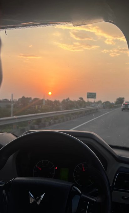 The image shows a car's interior with a view of an orange sunset through the windshield, creating a warm and serene atmosphere. The car appears to be stationary as the driver takes their eyes off the road for a moment. In the background, there are two cars on the highway, one closer to the foreground and another further away in the distance.