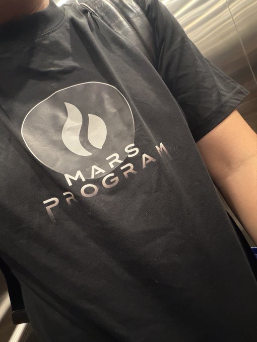 The image features a person wearing a black t-shirt with a white logo that says "mars program" on it. The person appears to be standing against a gray background, possibly indoors or at an event related to space exploration. The photo is taken from a low angle, making the person and their t-shirt the main focus of the image.