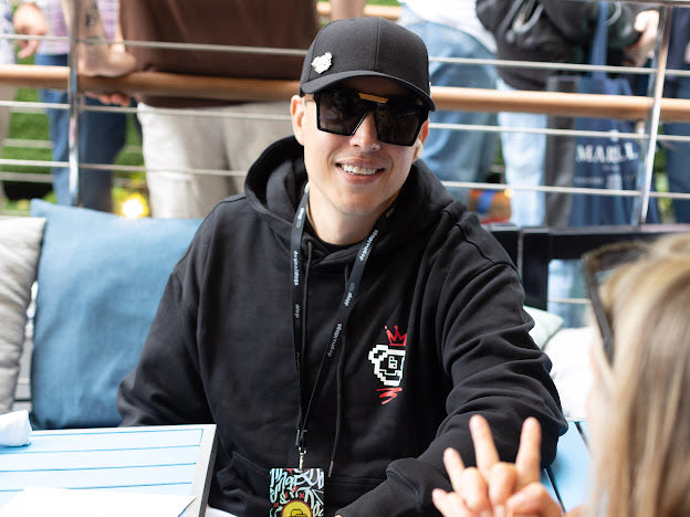 A smiling man wearing a black hoodie, sunglasses, and a baseball cap sits outdoors, making a peace sign with his hand.
