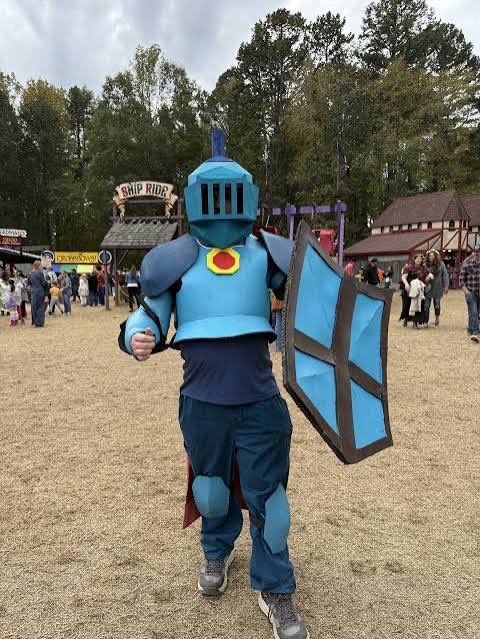 A person in a blue knight costume with a shield and helmet with a blue plume walks across a grassy area at a festival, with a "Skid Ride" sign visible in the background.