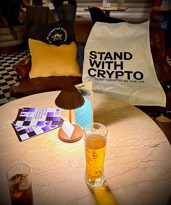 A casual photo of a cozy lounge area featuring a leather sofa with a "STAND WITH CRYPTO" pillow supporting crypto in the UK, along with a table displaying "Crypto Pub" flyers and a half-finished beer glass.