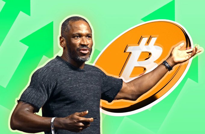 A man in a dark gray shirt gestures toward a large orange Bitcoin (₿) coin against a bright green background with upward arrows.