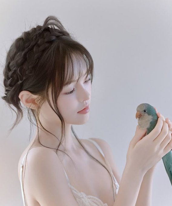 A young woman with braided hair gently holds a small green parrot while looking at it softly.