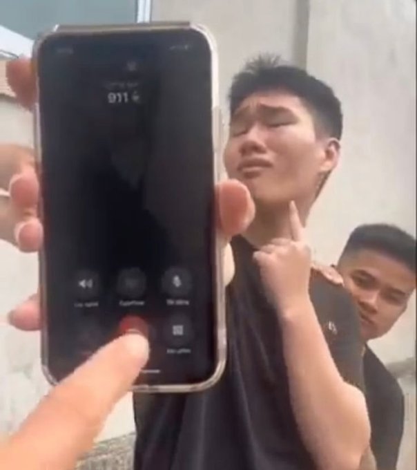 A young man crying with his fists clenched while someone holds up a smartphone showing an active phone call screen.