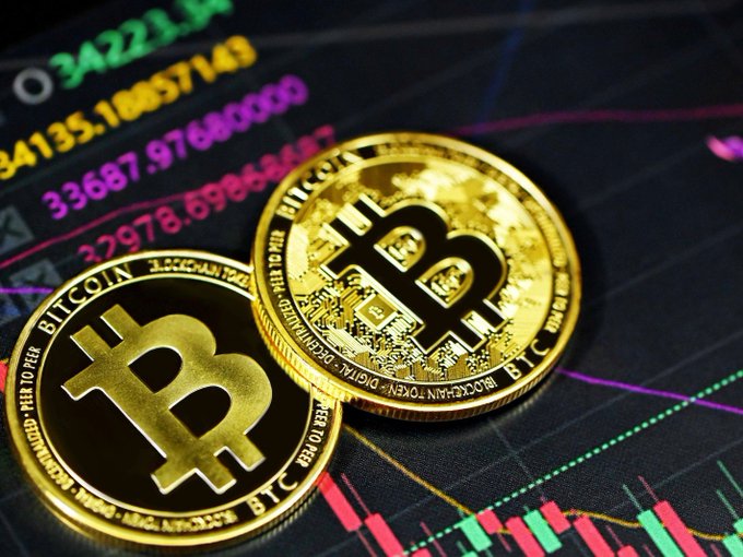 Two physical Bitcoin tokens rest on a stock chart display with visible numbers like 34221.14, 4135.18857149, 33687.976, and 27978.69, along with green and red candlestick patterns.