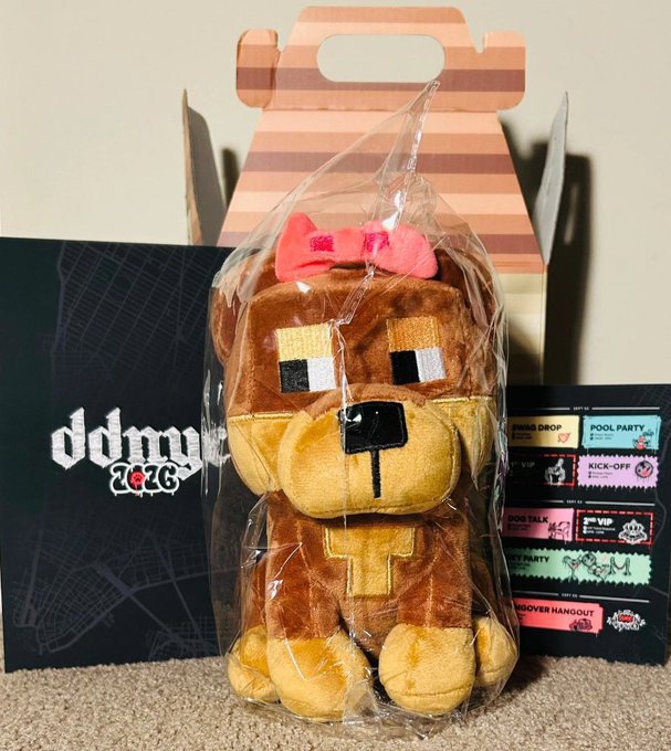 A packaged plush toy of a brown blocky bear with a pink bow tie, displayed with a 'ddmmw 2026' card and event tickets for 'POOL PARTY', 'KICK-OFF', and 'VIP'.