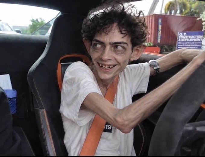A gaunt young man with curly hair and a wide grin grips a steering wheel inside a car, wearing a white shirt and an orange seatbelt with a "19" tag visible, while a container labeled "O.B. CONSTRUCTION & DEVELOPMENT" sits in the background. The image humorously illustrates the post's caption about the stress-free nature of memecoin trading, with the subject's exhausted, wild-eyed appearance poking fun at how emotionally and physically draining the volatile crypto market can actually be.
