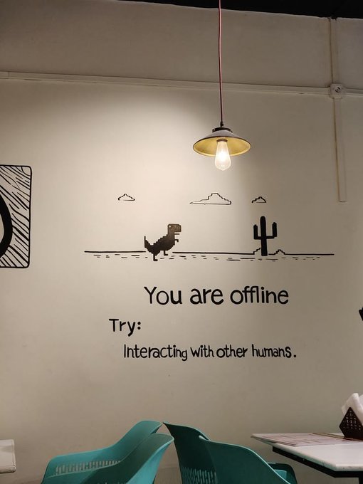 A café wall with a pixel-art dinosaur and cactus mural, displaying the ironic message 'You are offline. Try: Interacting with other humans.'