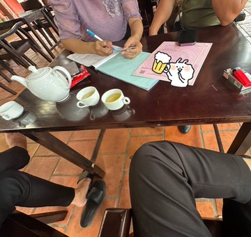 The image shows a person sitting at a table with a notebook and pen, possibly taking notes or working on a project. The table also has two cups of tea placed on it, suggesting that they might be enjoying a break during their work session. In addition to the person's feet visible in the foreground, there are other people present in the background, indicating that this scene takes place in a public setting like an office or café.