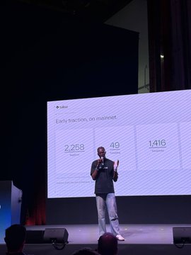 A man stands on stage giving a presentation in front of a large screen displaying the title 'Early traction, on mainnet' with three statistics: '2,258 Total Transactions', '49 Smart Contracts Deployed', and '1,416 Active Addresses'.